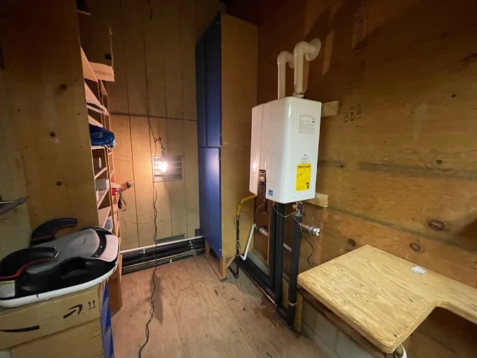 A modern white tankless water heater installed on a wood paneled wall in a residential storage or utility room.