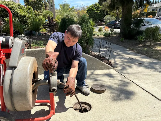 Plumbing technician wearing heavy duty protective gloves while operating a motorized drain snake to clear a main sewer line on a residential walkway.
