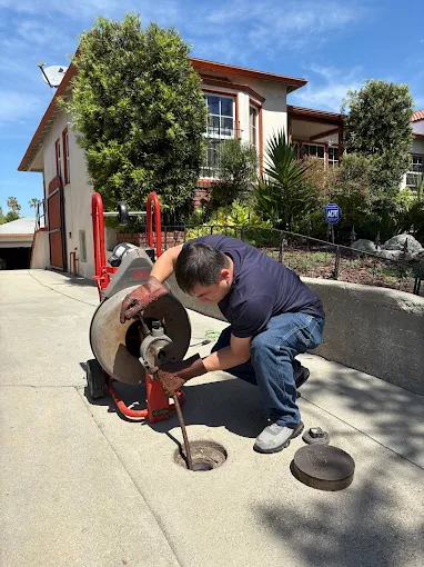 Plumbing technician wearing heavy duty protective gloves while operating a motorized drain auger to clear a main sewer line on a residential concrete driveway.