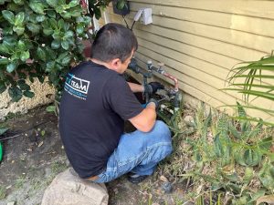 Plumbing technician wearing a black branded t-shirt and blue jeans while repairing exterior water lines near a residential house foundation.