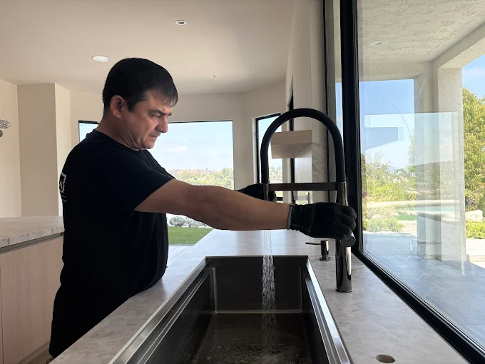 Plumbing technician installing a sleek matte black pull down faucet in a luxury kitchen with marble countertops and large windows.