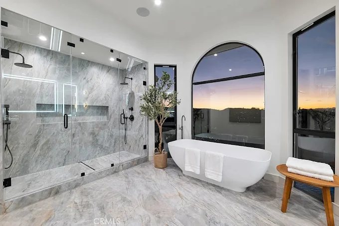 Luxury master bathroom with a large marble walk in shower and a white freestanding soaking tub in front of an arched window at sunset.