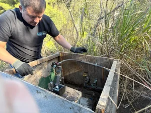 Plumbing technician wearing work gloves inspecting a water meter and backflow prevention assembly inside a wooden protective enclosure in a hillside area.