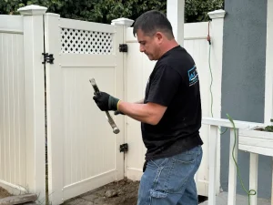 Plumbing technician in a black t-shirt and work gloves using a hammer to perform repairs on an outdoor fixture near a white vinyl fence.
