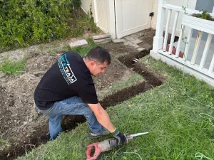 A plumbing technician from Art & Team uses a reciprocating saw to cut pipes inside a freshly dug trench for a residential yard drainage system.