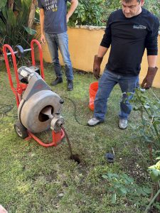 A plumbing technician using a heavy duty motorized drain auger to clear a blockage in an outdoor residential sewer line.