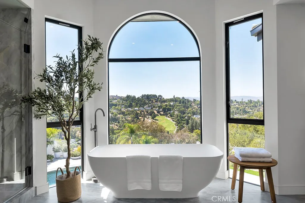 Modern white freestanding soaking tub positioned in front of a large arched window overlooking a scenic green valley in a luxury master bathroom.