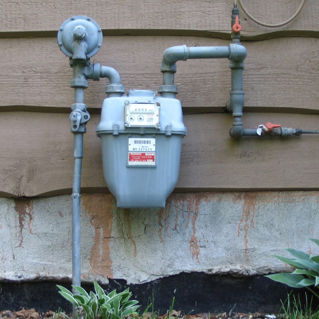 A residential gas meter located outside of a house with a wall behind it.