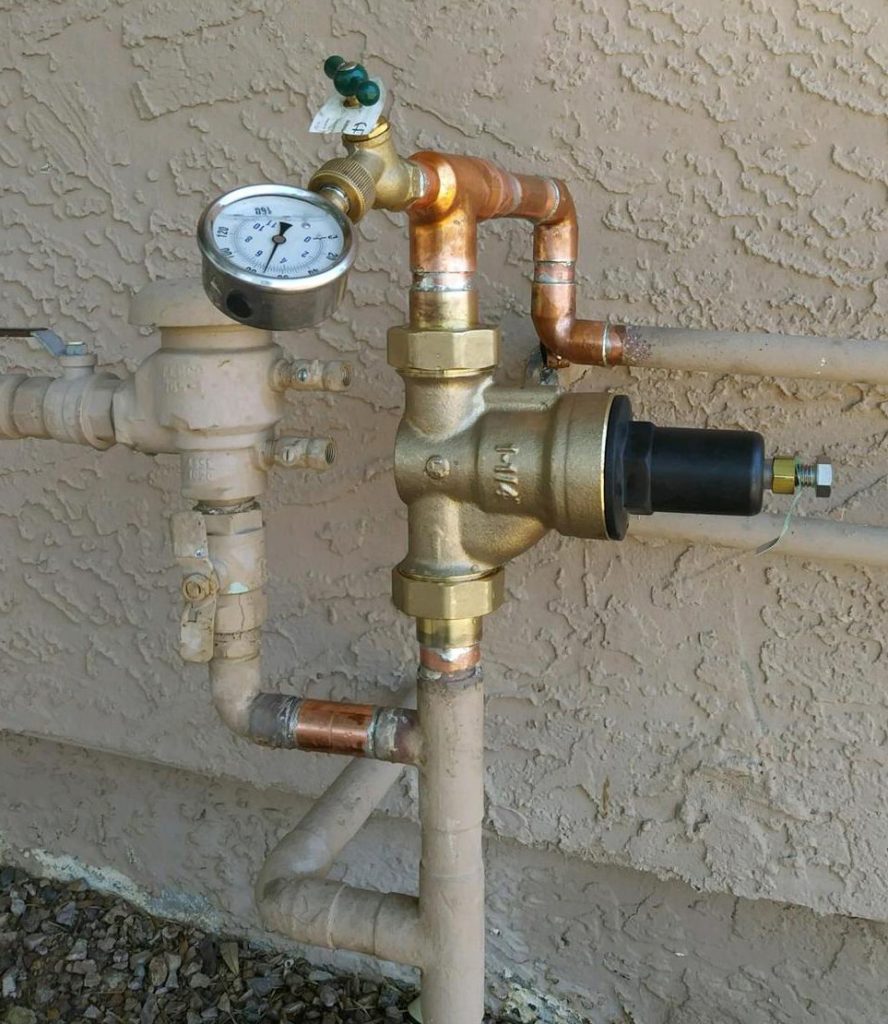Exterior water pressure regulator valve with a circular pressure gauge installed on a copper main line.