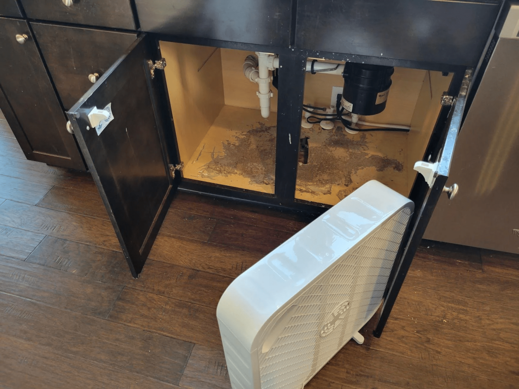 The kitchen cabinets are open and a fan is drying the cabinet after a water leak damaged the wood.