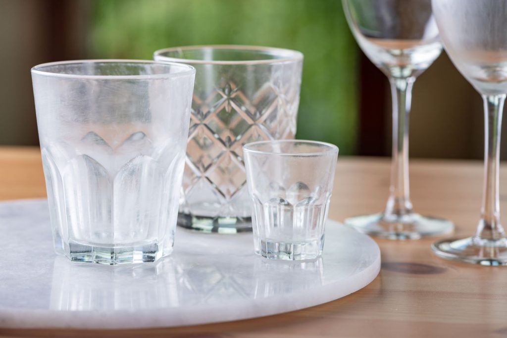 Four assorted drinking glasses and wine glasses with a cloudy white film from hard water mineral buildup sitting on a marble tray.
