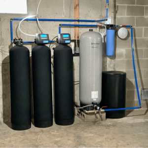 Professional basement installation of a multi stage water filtration system featuring three black carbon tanks and a large grey Pentair storage tank connected with blue PEX piping.