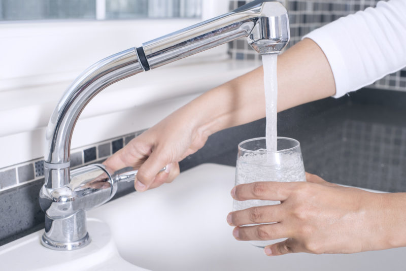 Close up of a person filling a clear glass with fresh, filtered water from a modern chrome kitchen faucet.