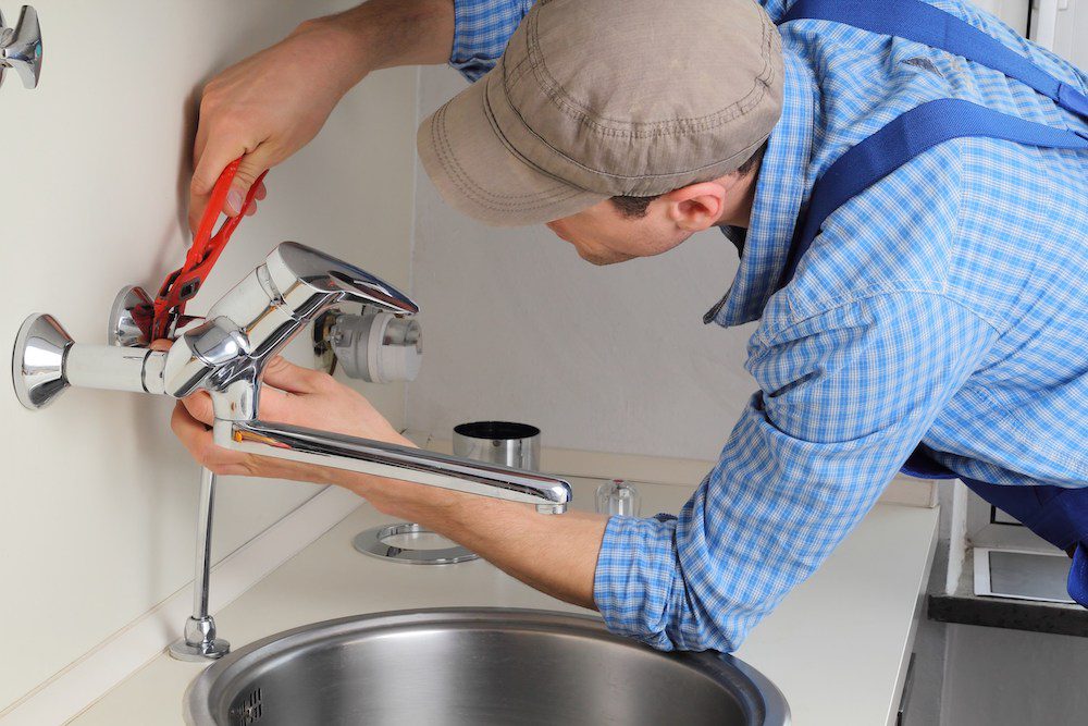 A man installing a wall mounted chrome lavatory faucet and tightening it with tongue and groove pliers.
