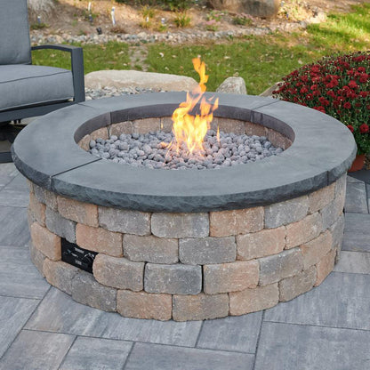 Gas fire pit with stoned walls and an active fire in the backyard of a home with the lawn and a chair in the background.