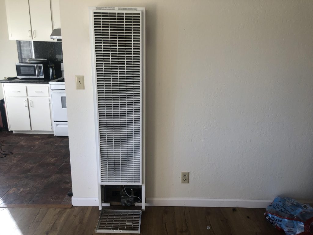Outdated wall heater mounted on a white wall near the kitchen with the service panel open.