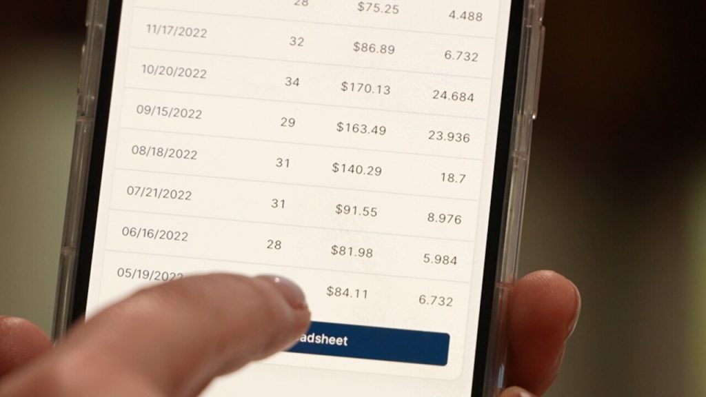 A person using a smartphone to review a monthly utility billing spreadsheet with rising costs and water usage data.