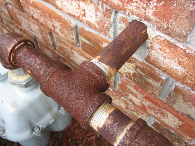 Very rusted galvanized gas pipe going into a brick wall and is connected to a gas meter below the pipe.