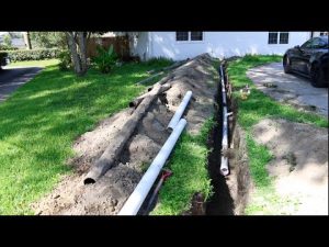 A long trench in a residential yard showing the installation of a new white PVC sewer pipe.