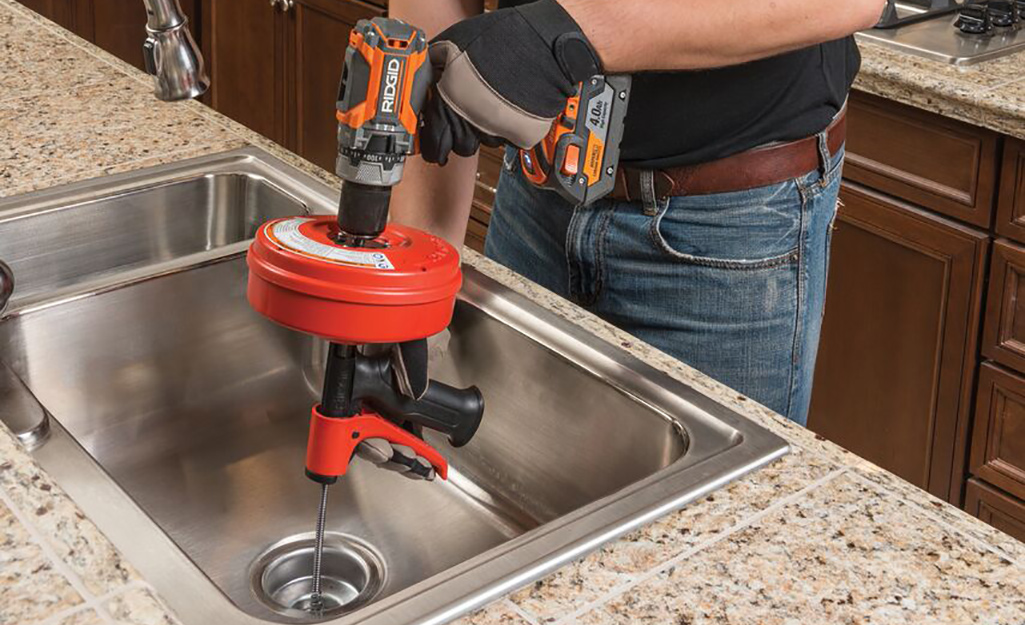 A man snaking a kitchen sink with a handheld snake powered by a cordless drill.