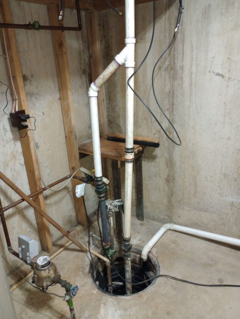 A wide shot of a sump pump pit located in the basement of a home with drainage pipes going into and out of the pit.