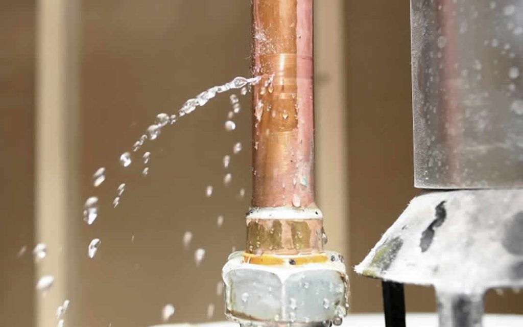 Close up of a high pressure pinhole leak spraying water from a vertical copper pipe joint.