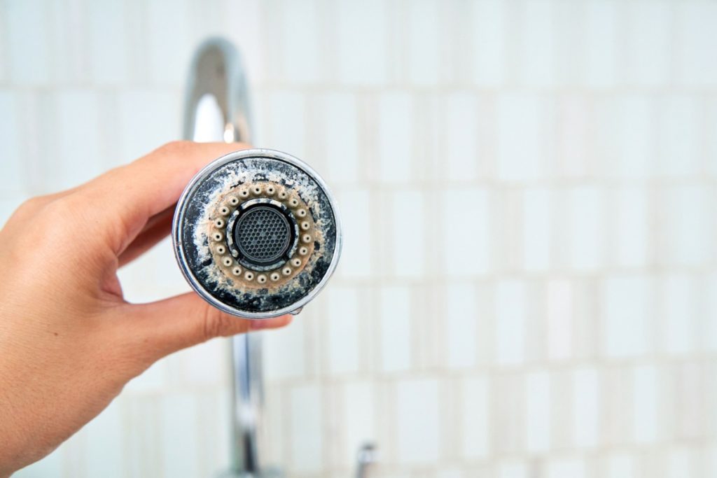 Hand holding a chrome shower head covered in thick white calcium and lime scale deposits from hard water.