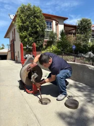 Plumbing technician wearing heavy duty protective gloves while operating a motorized drain auger to clear a main sewer line on a residential concrete driveway.