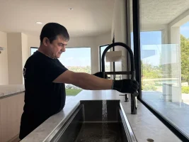 Plumbing technician installing a sleek matte black pull down faucet in a luxury kitchen with marble countertops and large windows.