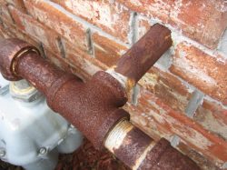 Very rusted galvanized gas pipe going into a brick wall and is connected to a gas meter below the pipe.